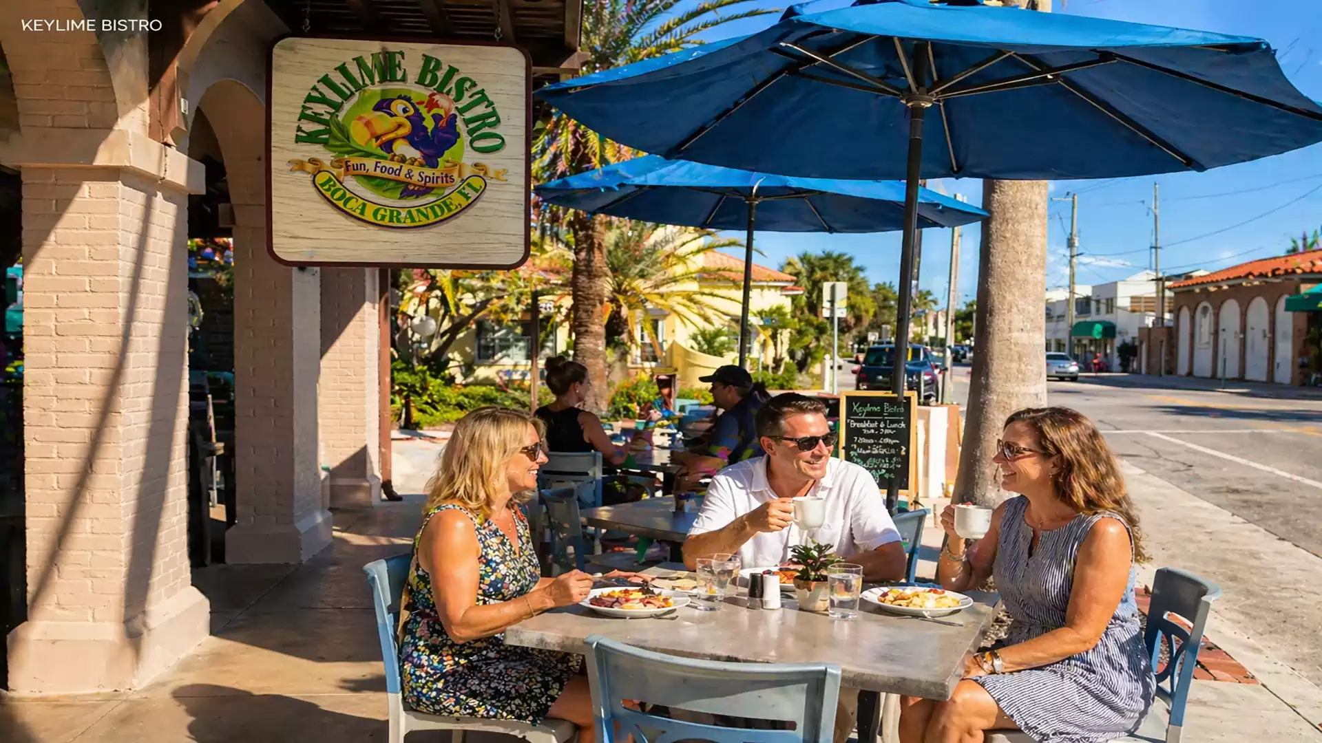 Three people enjoying an outdoor lunch under a blue umbrella in front of a restaurant called Keylime Bistro. They are seated at a table on a sunny street.