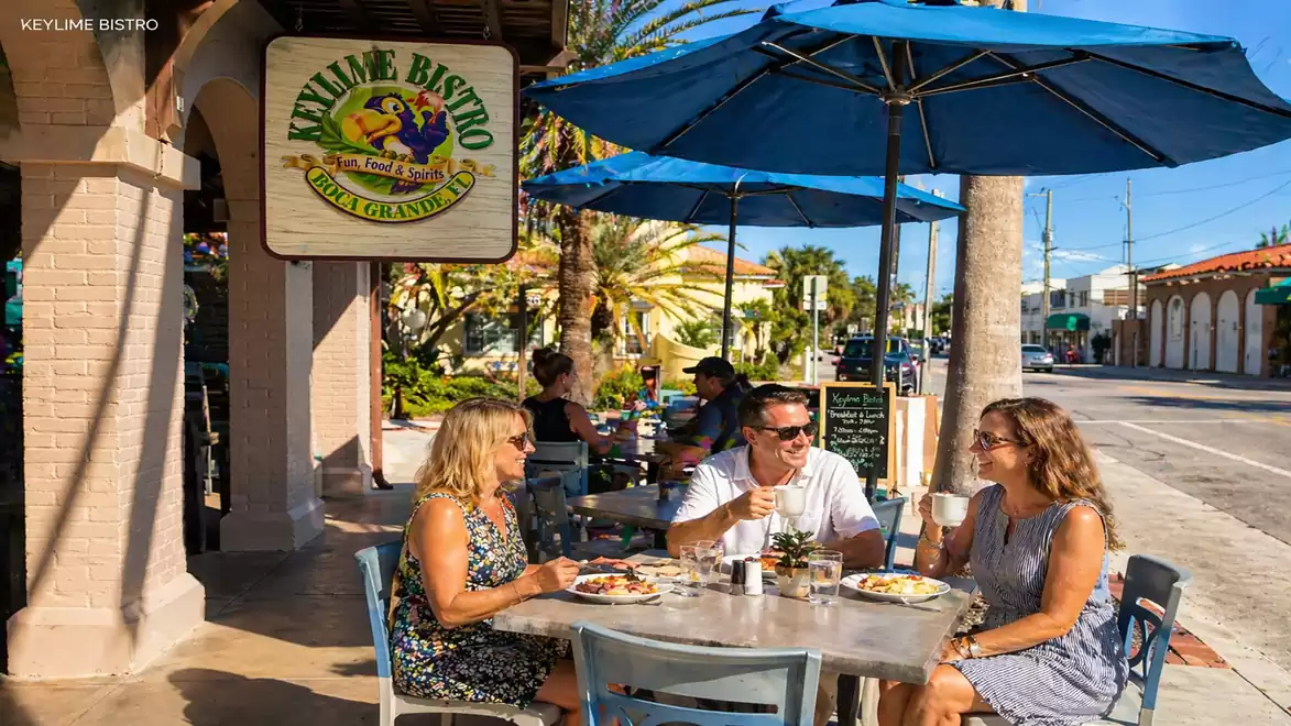 Three people enjoying an outdoor lunch under a blue umbrella in front of a restaurant called Keylime Bistro. They are seated at a table on a sunny street.