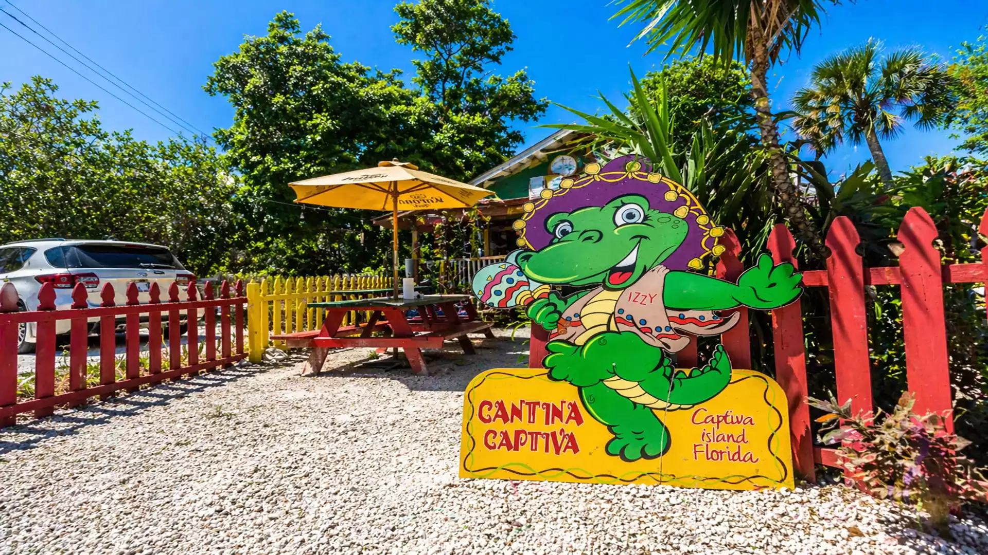 Colorful restaurant with a cheerful cartoon alligator sign in a garden with gravel ground, red fence, and parasol on Captiva Island, Florida.