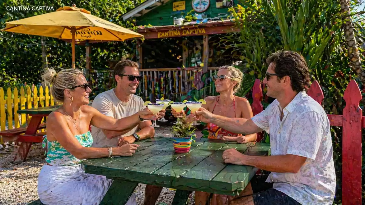 Four people enjoying cocktails at an outdoor table with a colorful restaurant in the background.