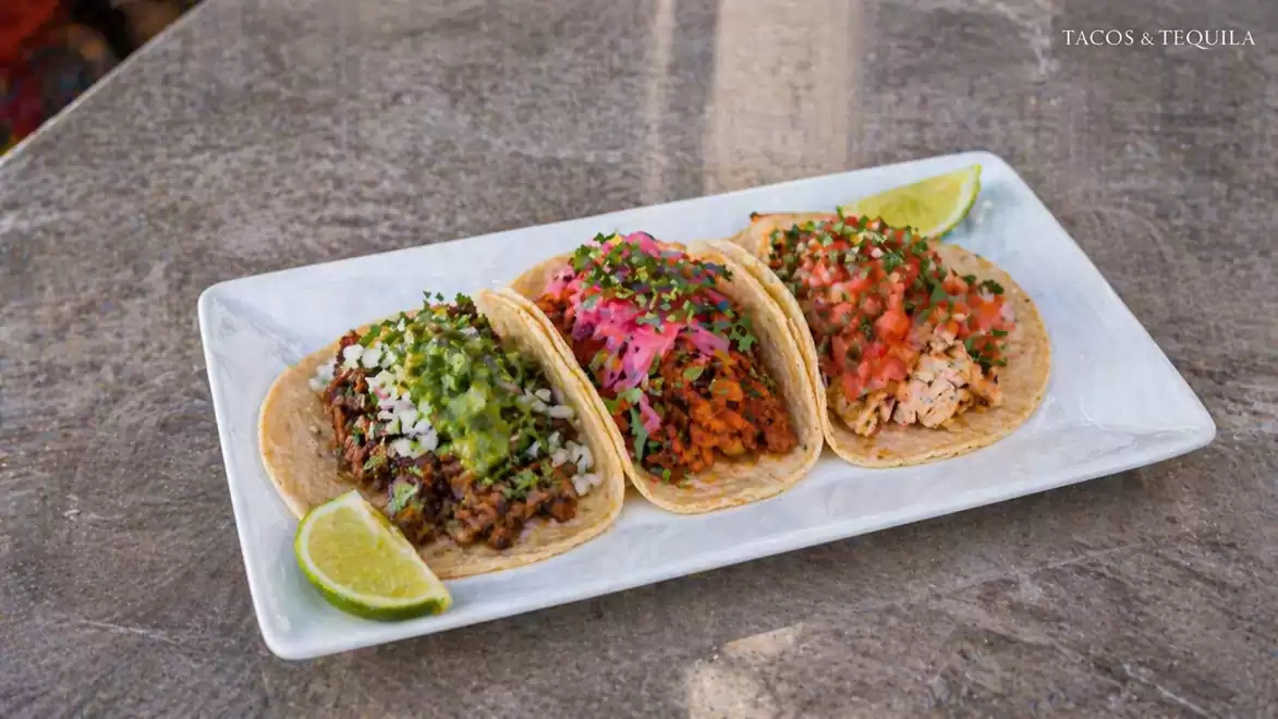 Three delicious tacos served on a rectangular white plate with lime slices, filled with various fresh ingredients.