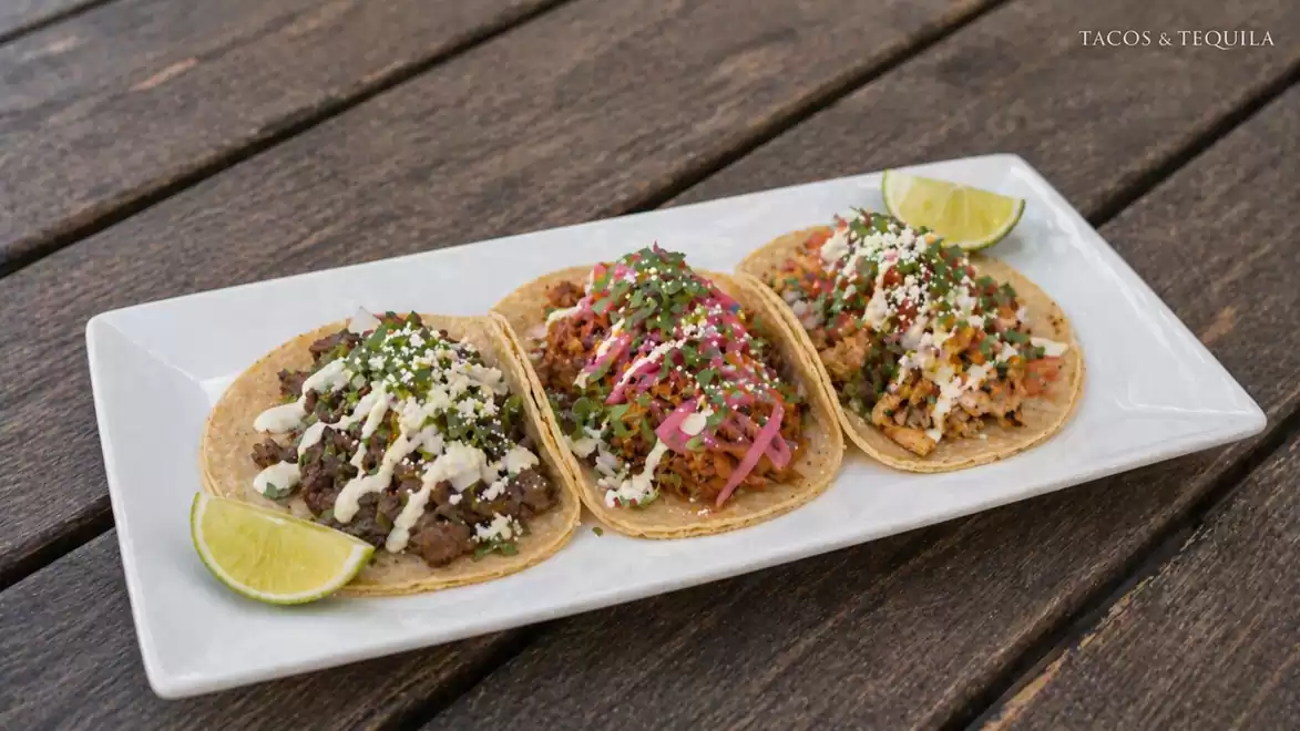 Three tacos with different toppings on a white plate, garnished with lime wedges.