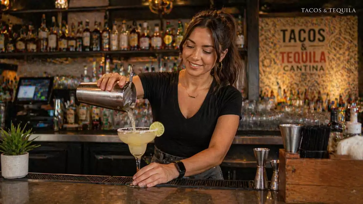 A bartender pours a cocktail in a lively bar setting. In the background, there are shelves with bottles and a sign reading 'Tacos & Tequila Cantina'.