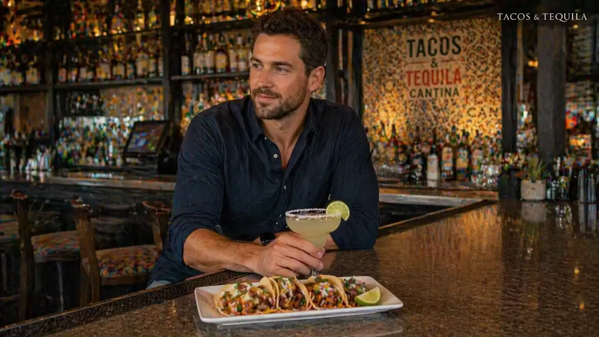 A man sits at a bar in front of a plate of tacos and a glass of margarita. The bar is stocked with bottles in the background.