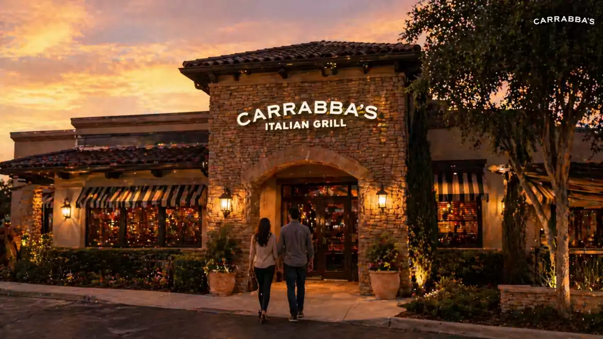 A couple enters Carrabba's Italian Grill at sunset. The restaurant features a rustic stone facade with warm lighting and is surrounded by trees.