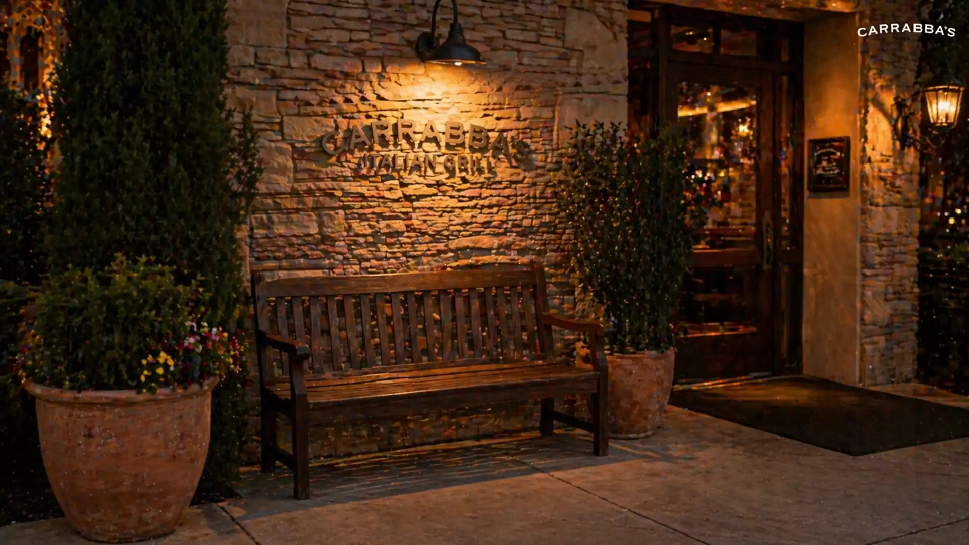 Evening view of a cozy restaurant entrance with stone wall, wooden bench, and plants.