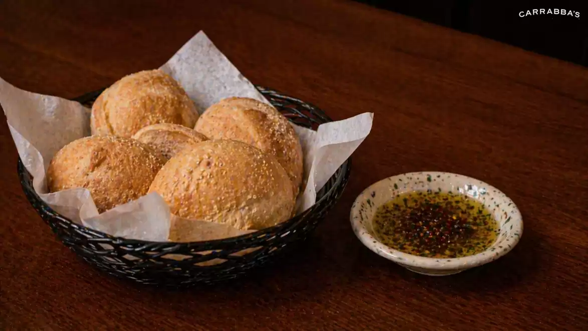 A basket of fresh bread rolls on a wooden table, next to a plate with olive oil and spices.