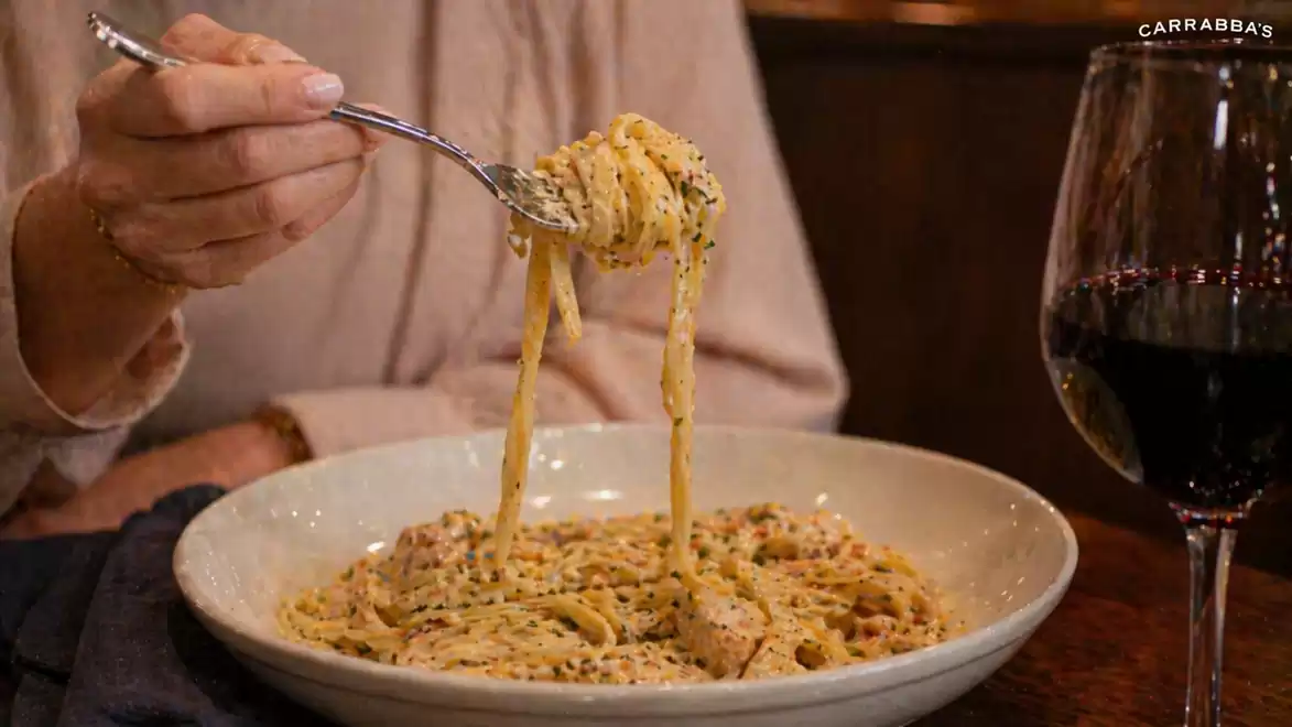 A person lifts a portion of spaghetti with creamy sauce from a white plate using a fork. A glass of red wine is placed beside it.