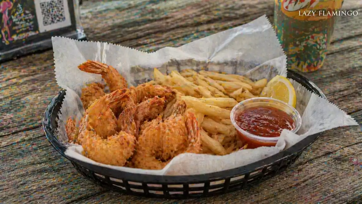 Coconut shrimp with fries and cocktail sauce served in a black basket, alongside a drink in a plastic cup.