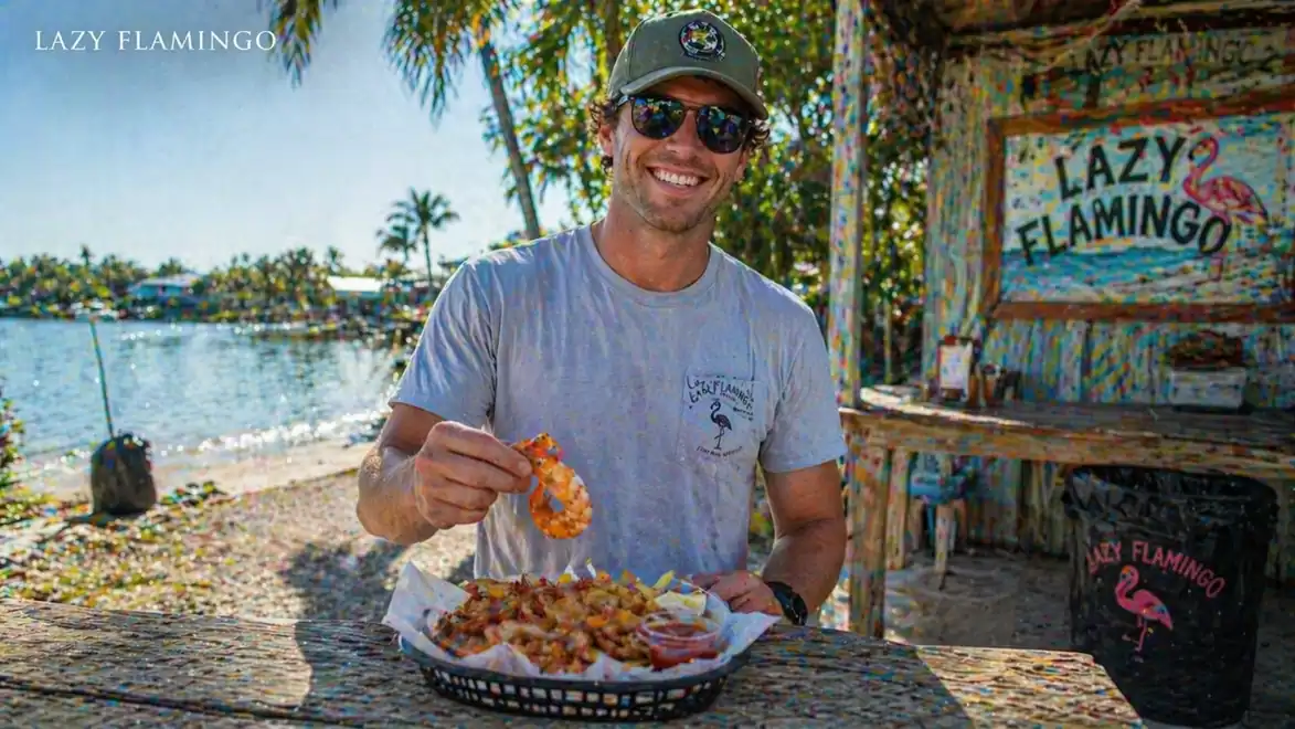 A man is smiling while sitting in front of an outdoor table at Lazy Flamingo. He is holding a shrimp dish. In the background, there is a picturesque beach setting with palm trees.