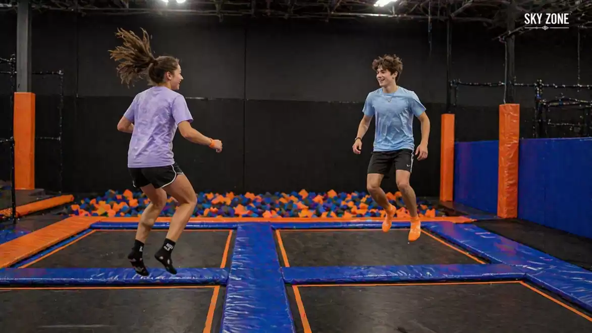 Two people jumping on trampolines at an indoor sports facility with a foam pit in the background.