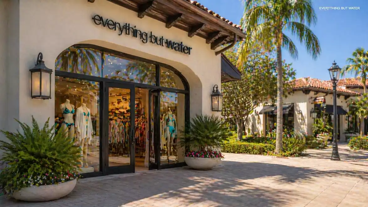 A fashion store with large windows displays trendy clothing and accessories amidst palm trees in sunny weather.