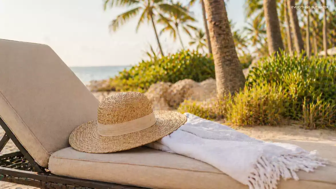 A comfortable lounge chair on the beach with a straw hat and a white towel, surrounded by palm trees and green vegetation, overlooking the sea.