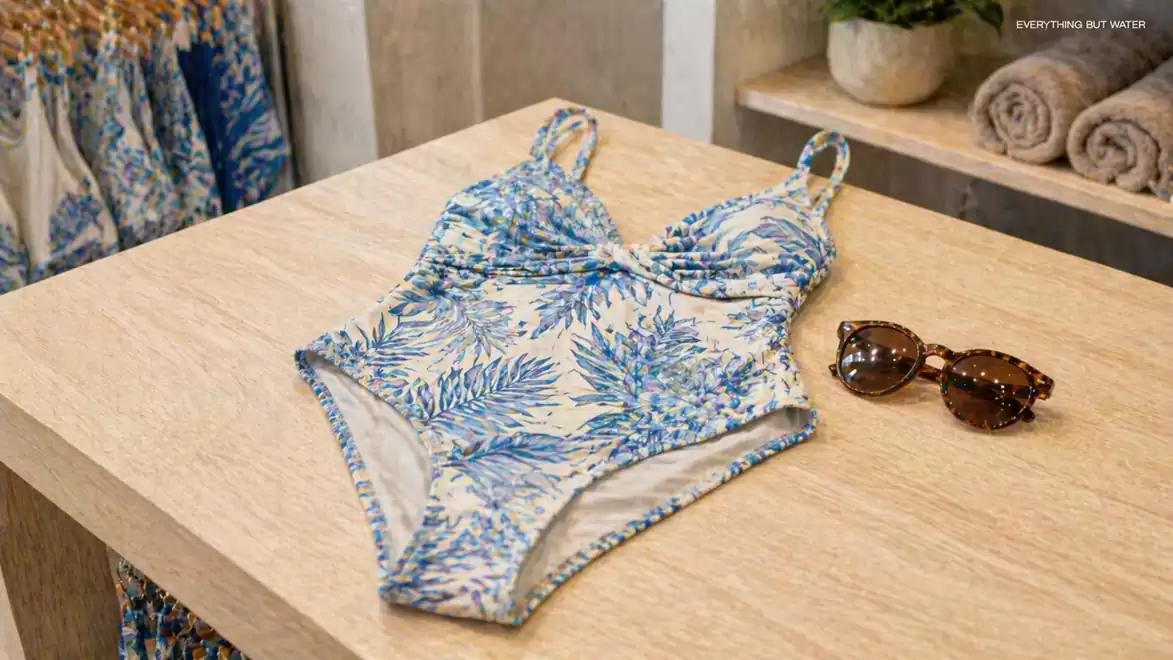 A blue and beige swimsuit with a tropical leaf pattern lies on a wooden table next to sunglasses. Towels and a plant are visible in the background.
