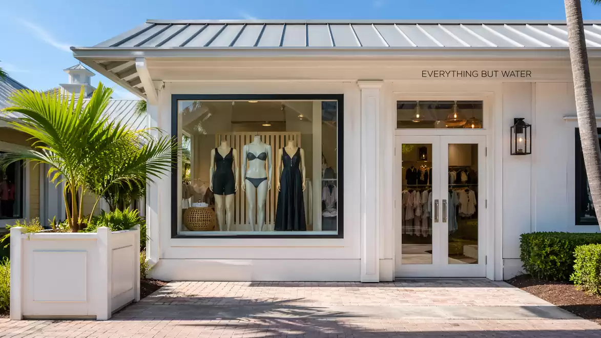 Exterior view of a stylish clothing store with swimsuits in the window, surrounded by neatly trimmed plants.