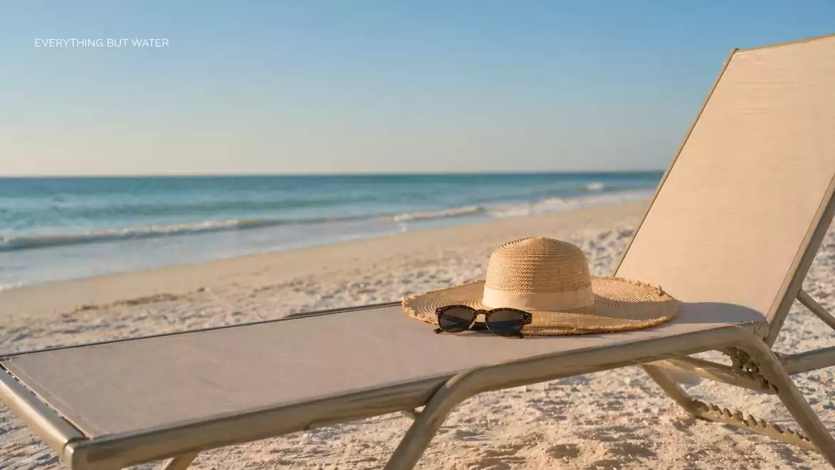 A lounge chair with a straw hat and sunglasses is on a sunny beach overlooking the sea.