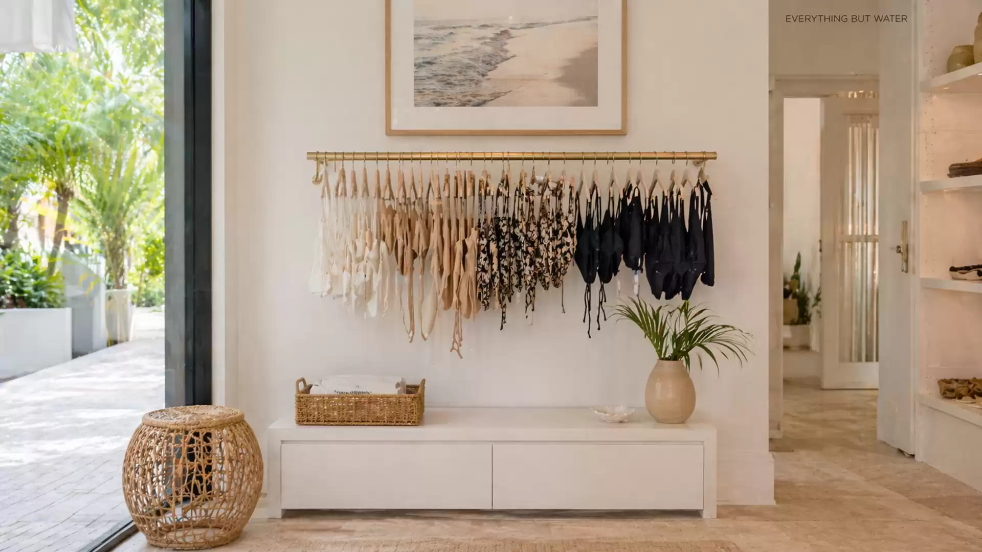 Elegant room with light walls, a small side table, and a collection of bikinis on a rack. Large glass windows offer a view of a green outdoor area.