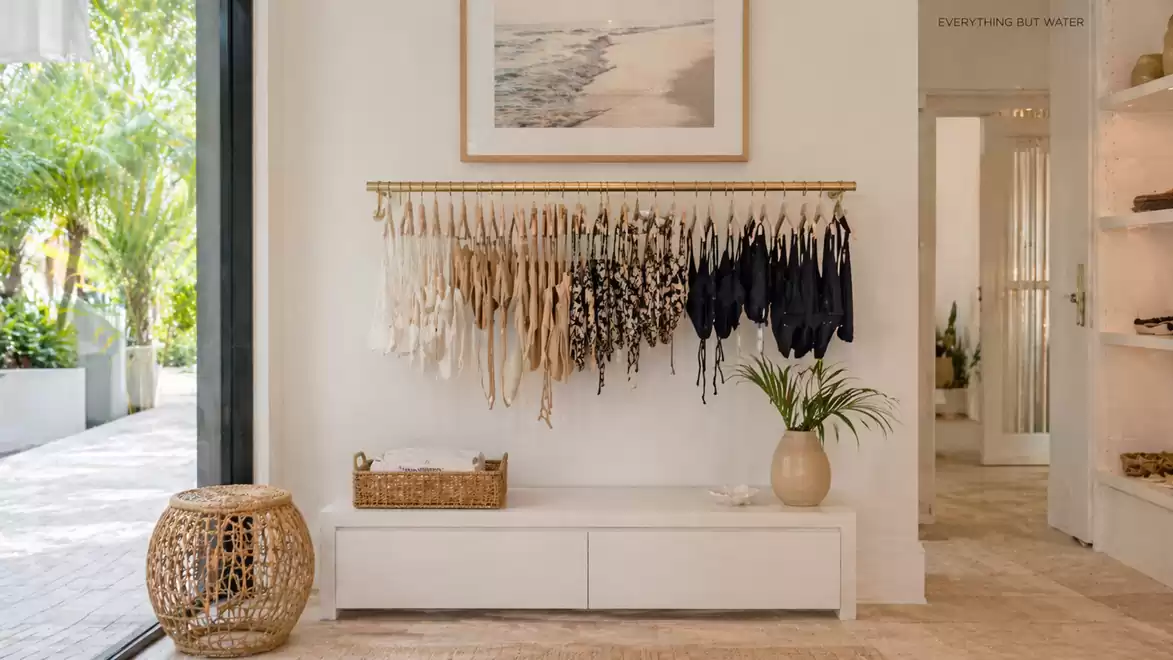 Elegant room with light walls, a small side table, and a collection of bikinis on a rack. Large glass windows offer a view of a green outdoor area.