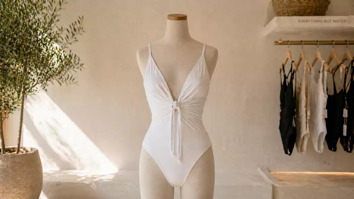 Elegant white swimsuit on a mannequin in a modern, minimalist room with plants and hanging clothes.