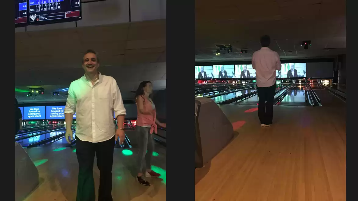 Two people at a bowling alley, one person smiling at the camera and another preparing to throw a ball.