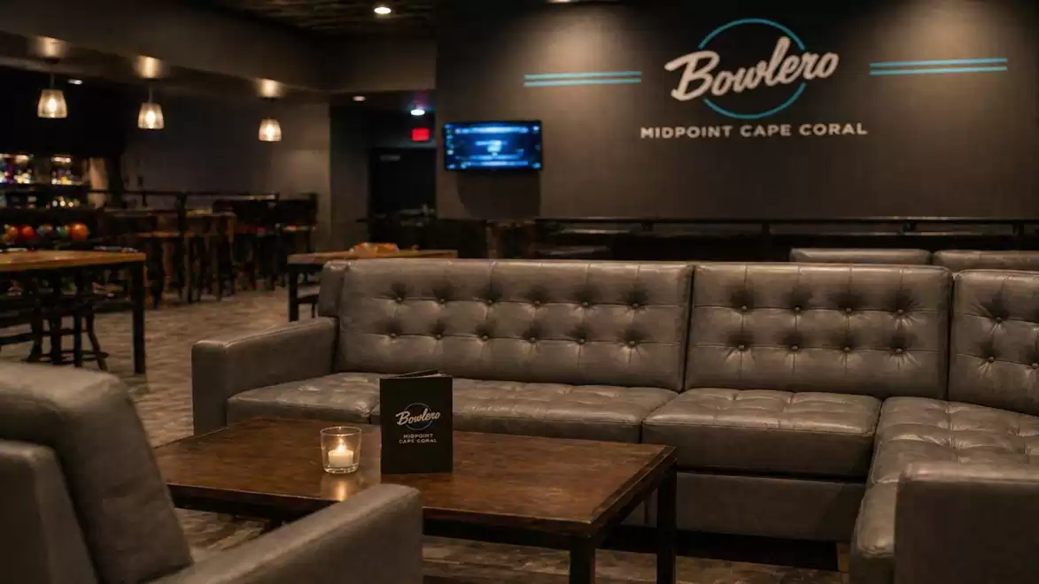 Modern lounge with leather sofas and a table, in the background a bar with the 'Bowlero Midpoint Cape Coral' logo on the wall.