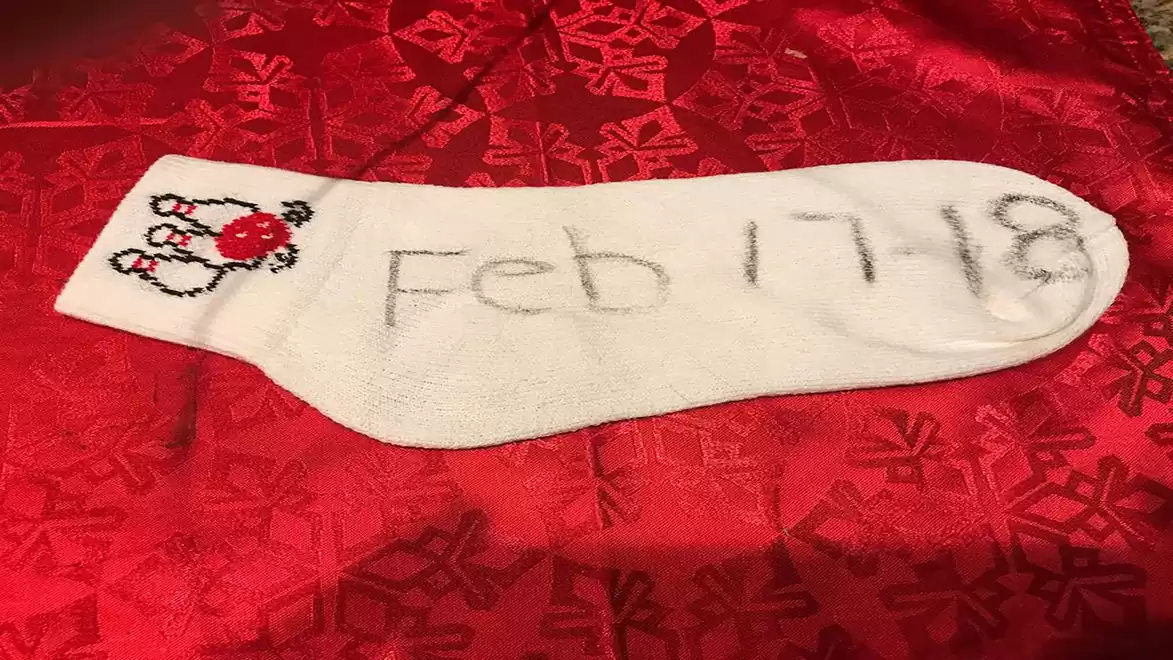 A white sock with the red and white date 'Feb 17 18' on a red tablecloth with snowflake pattern.