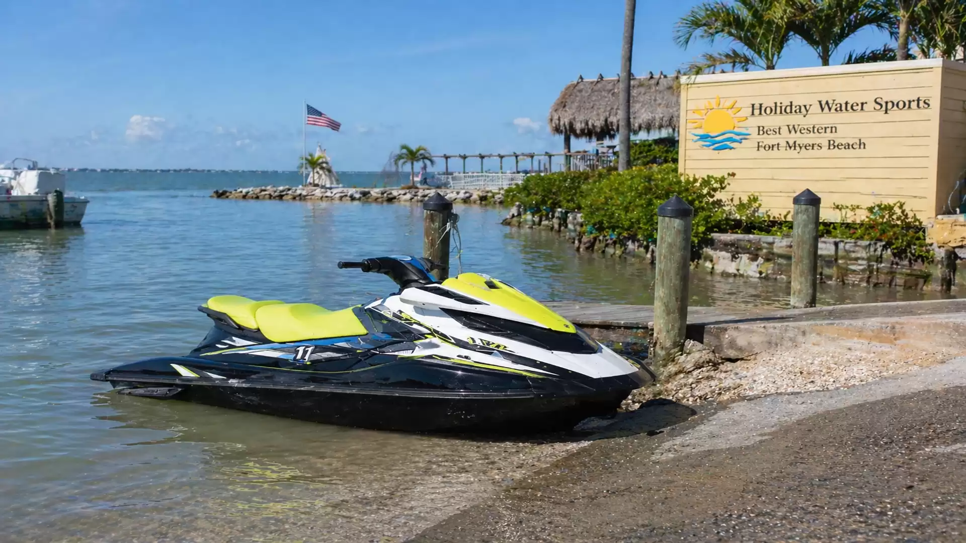 A green jet ski on the shore of a calm lake, with a Holiday Water Sports sign nearby and palm trees in the background.