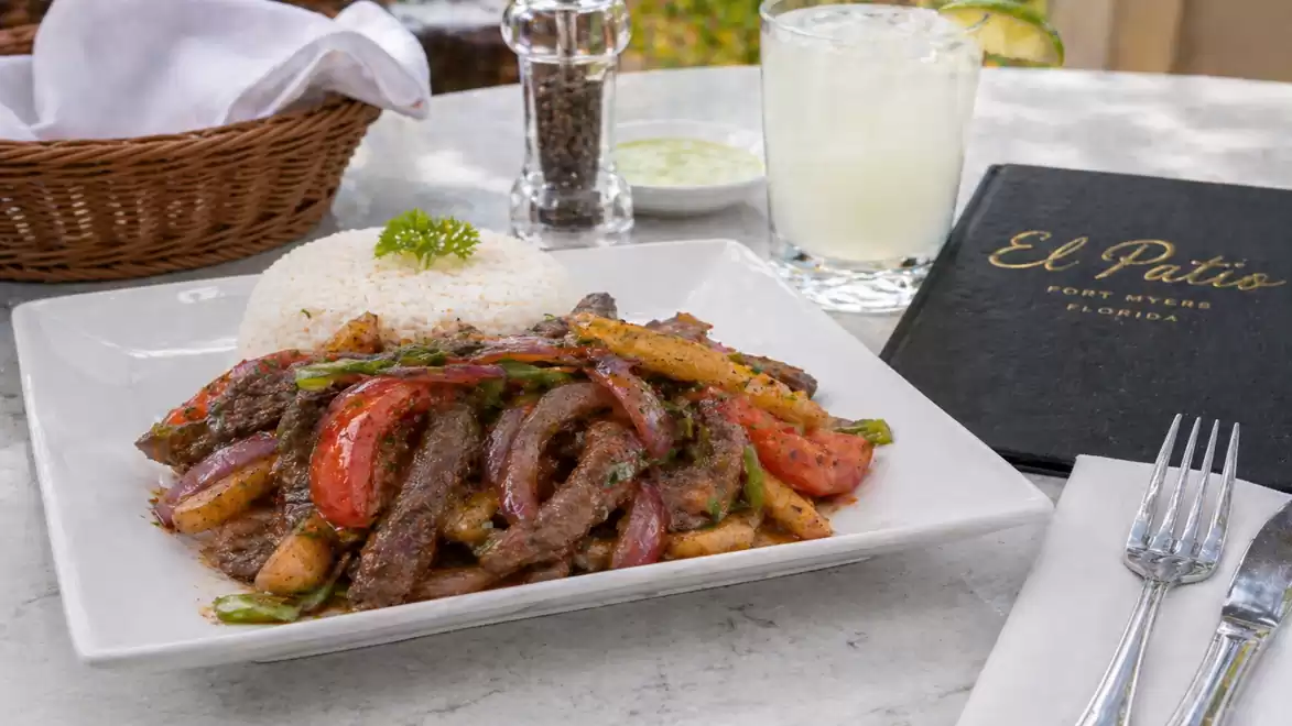 A plate of Lomo Saltado, a Peruvian dish with beef, tomatoes, onions, and peppers, served with rice on a table with a drink in the background.