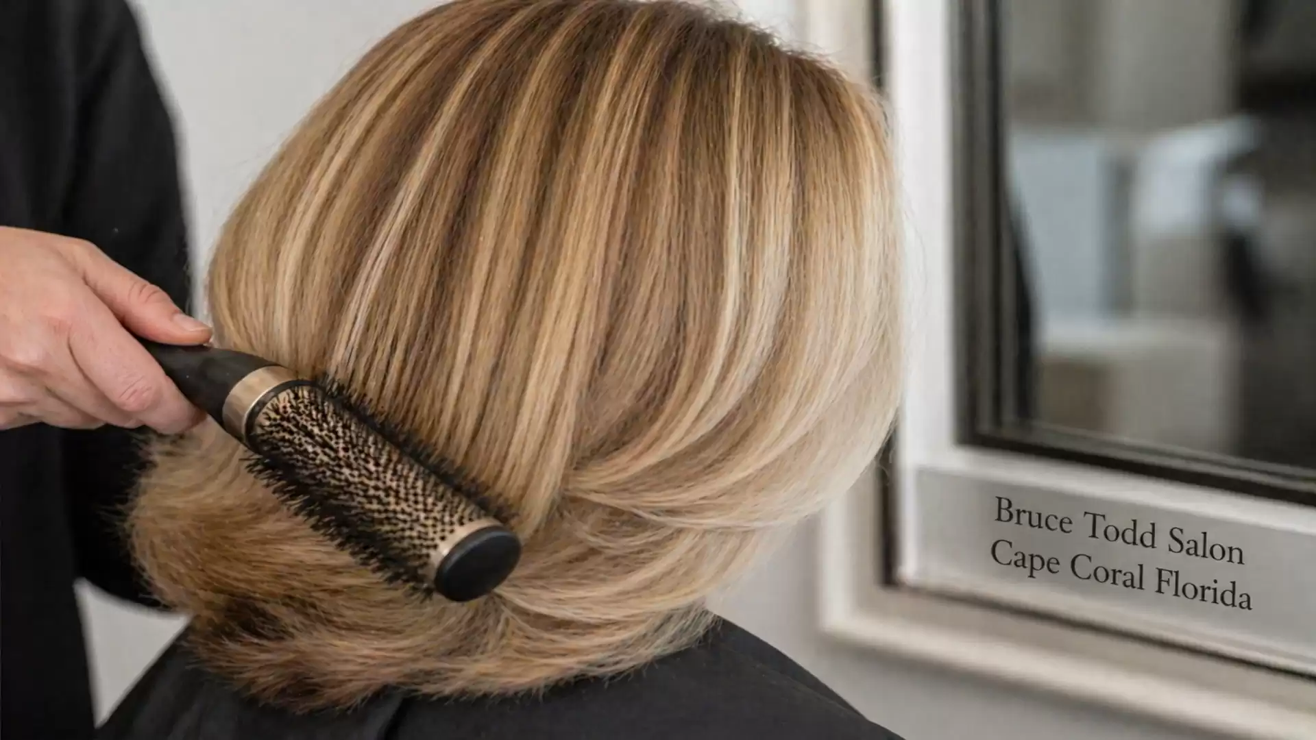 A person in a hair salon is getting styled with a round brush. In the mirror, the phrase 'Bruce Todd Salon, Cape Coral Florida' is visible.