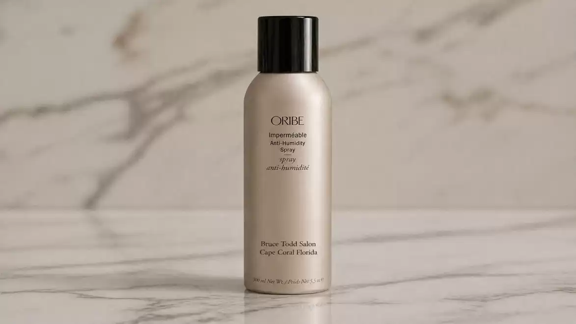 A bottle of texturizing spray with a black cap is placed on a marble surface against a blurred background.