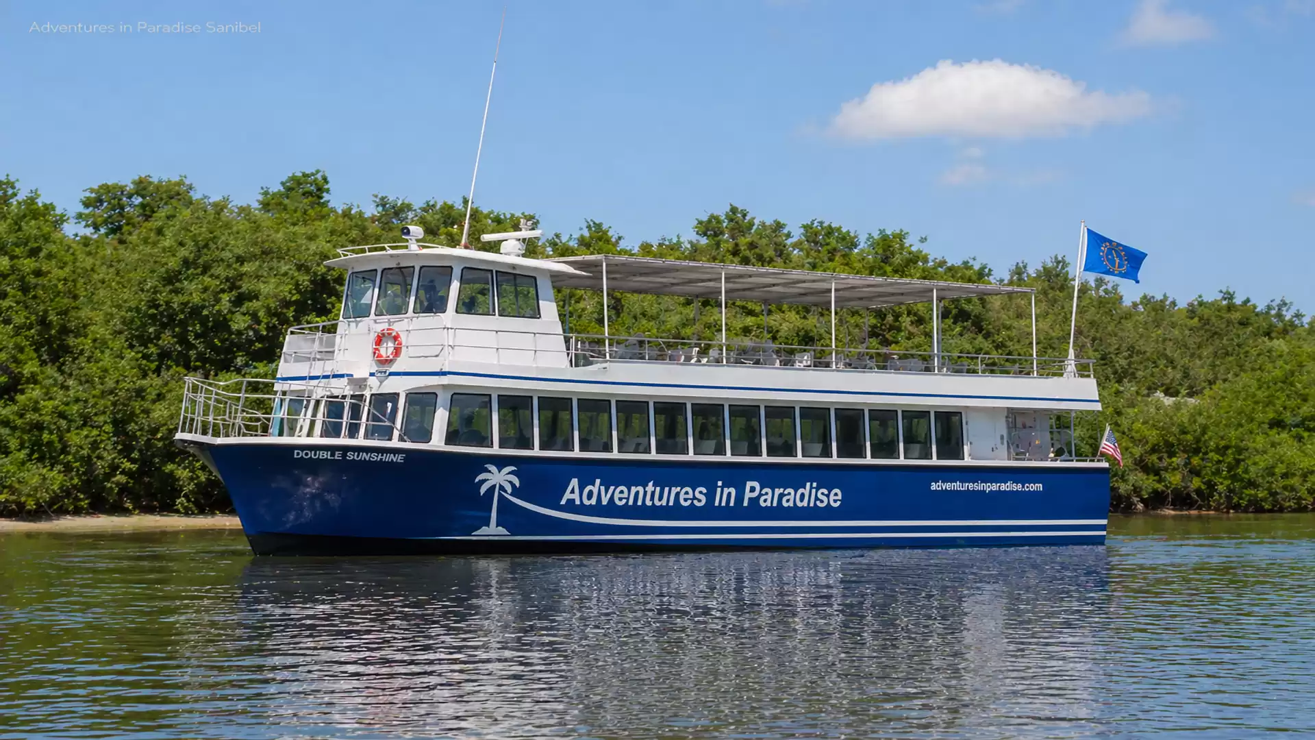A blue and white excursion boat on a calm river with the text 'Adventures in Paradise', surrounded by green vegetation and a blue sky.