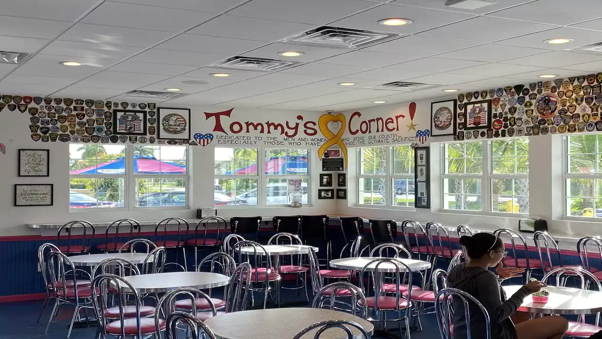A diner with a sign above 'Tommy's Corner'. The room has many tables and chairs, along with decorative plaques and photos on the walls. A person is sitting at a table.