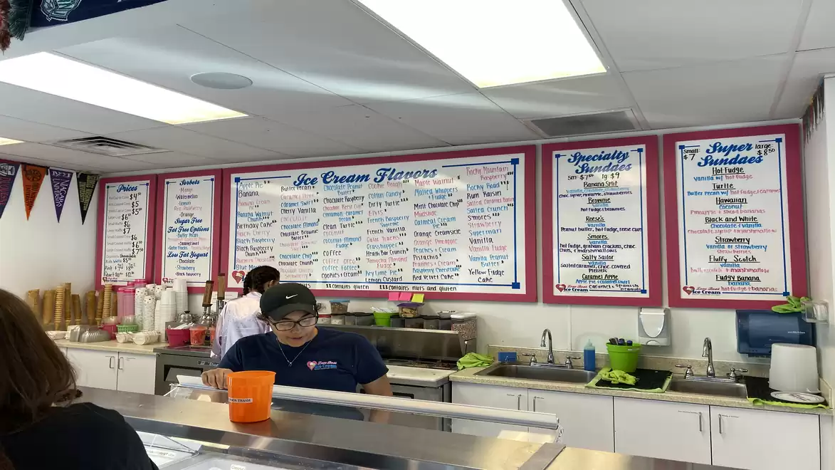 An ice cream shop with employees behind the counter in front of a large board showing ice cream flavors and specialties.