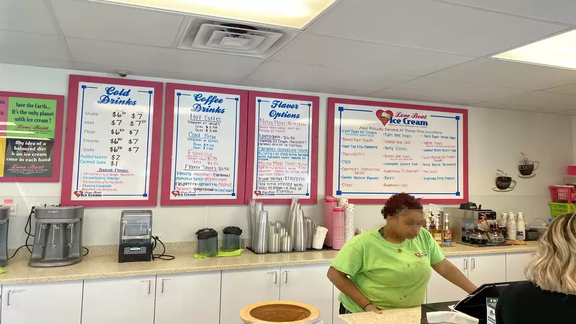 Interior of an ice cream shop with two people. One person is standing behind the counter. Menus with drinks and ice cream flavors are on the wall.