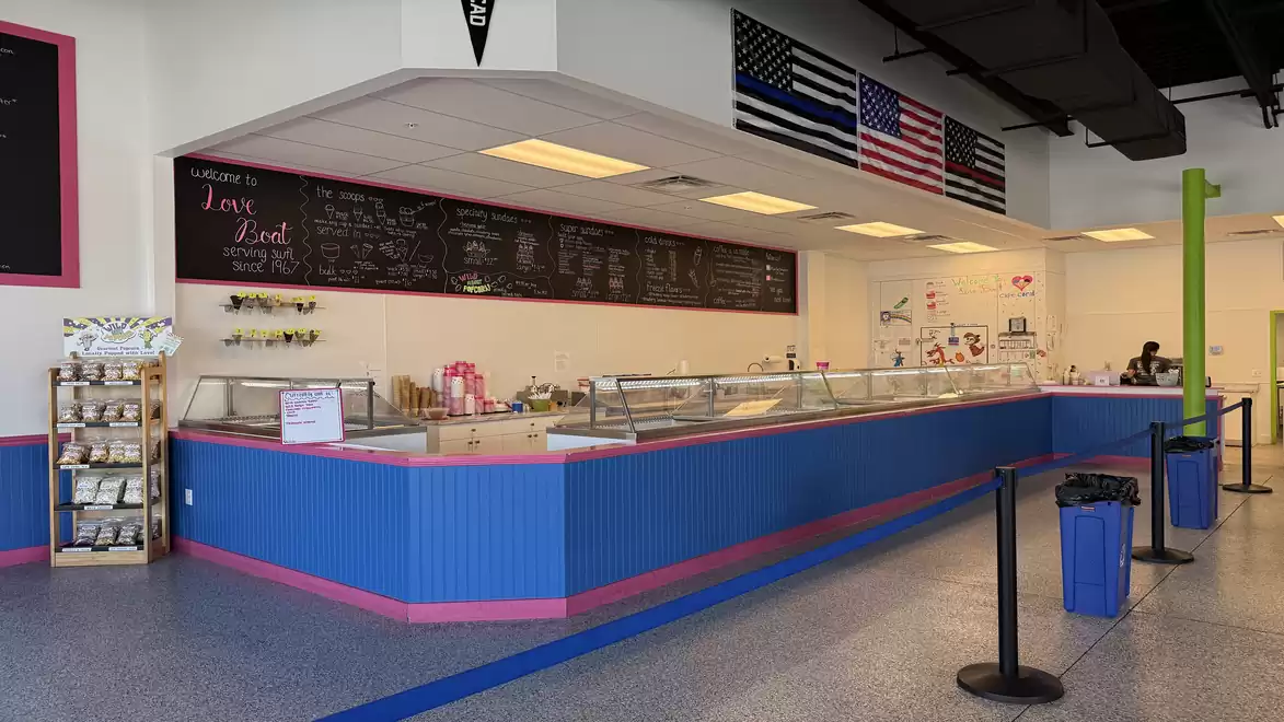 Interior of an ice cream shop with a counter painted in blue and pink, and a blackboard menu. Flags are hanging on the walls. The floor is gray, and there are trash bins near the counter.