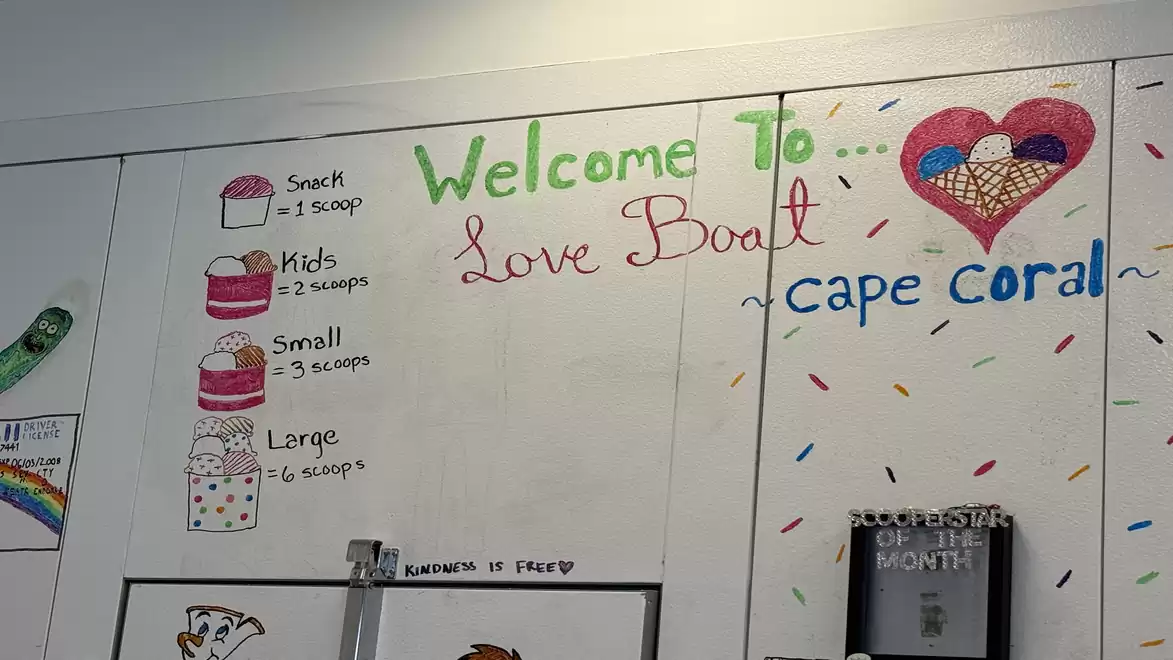 Interior view of an ice cream shop with a painted board showing ice cream portions and prices. Various sizes range from Snack to Large. Welcome message 'Welcome to Love Boat' on the wall.