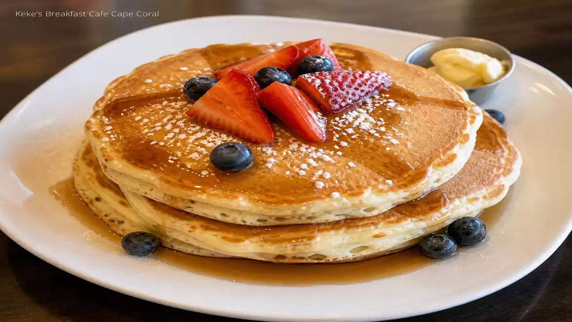 A plate with two large pancakes topped with strawberries and blueberries, sprinkled with powdered sugar, with a small dish of butter on the side.
