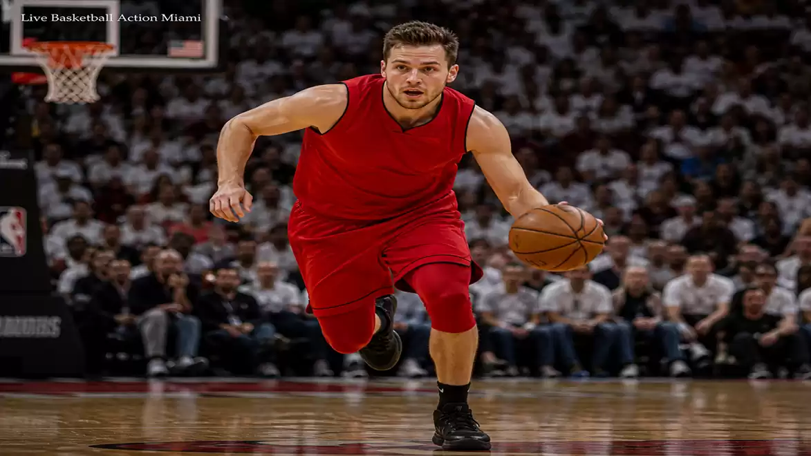 A basketball player in a red jersey is dribbling on the court during a game. Blurred spectators are visible in the background.