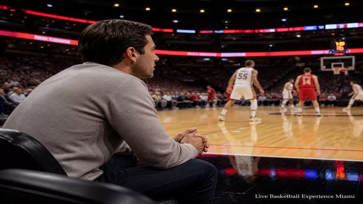 A man sits courtside during a basketball game in a large arena, while players in red and white actively engage on the court.