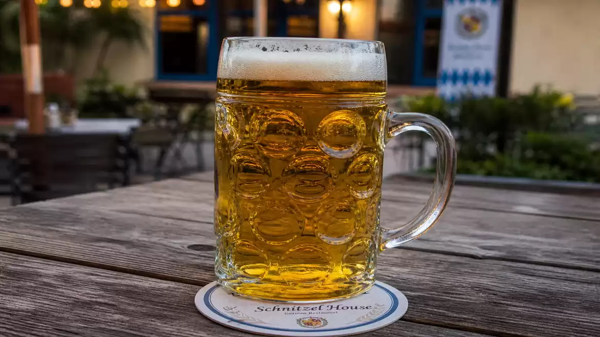 A traditional beer mug with golden beer on a wooden table outdoors, surrounded by a beer garden atmosphere.