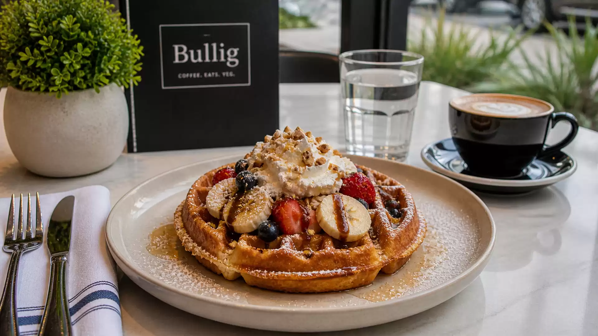 A deliciously presented plate with a waffle topped with fresh fruits and cream is on a table next to a cup of coffee at Bullig Café.