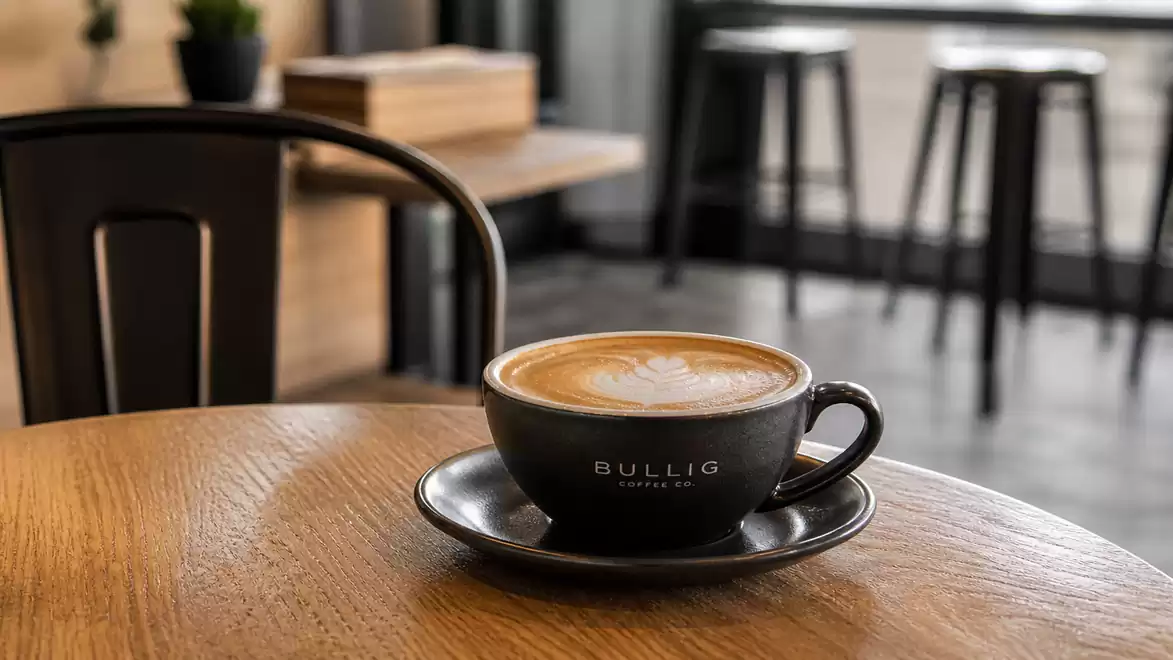 A cup of cappuccino on a wooden table in a modern café with blurred barstools in the background.
