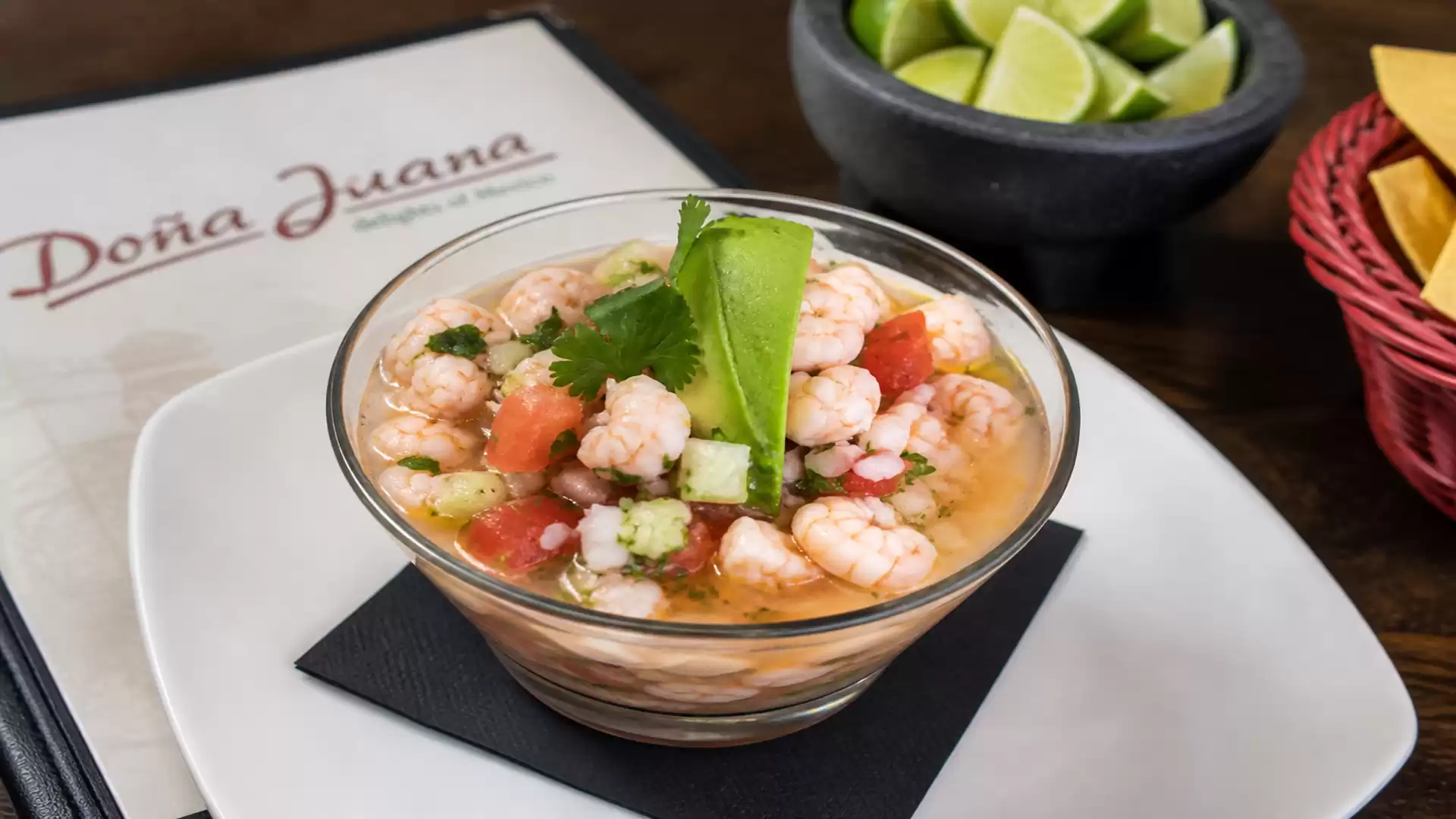 A bowl of ceviche is on a table next to a menu. The dish contains shrimp with avocado and cilantro, served with lime wedges and tortilla chips.