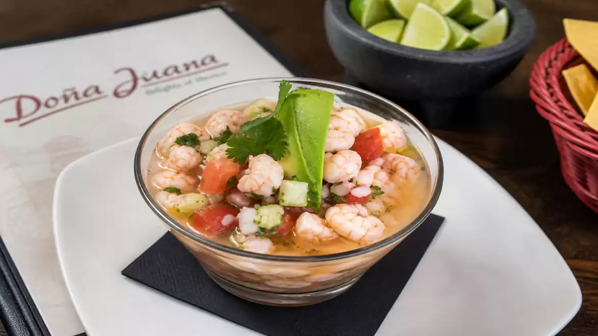 A bowl of ceviche is on a table next to a menu. The dish contains shrimp with avocado and cilantro, served with lime wedges and tortilla chips.