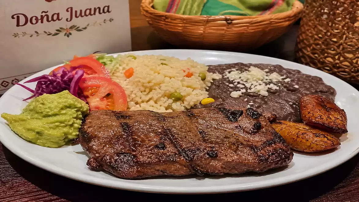 A plate with grilled steak, rice, beans, fried plantains, and sides like guacamole, tomatoes, and coleslaw at Restaurant Dona Juana.