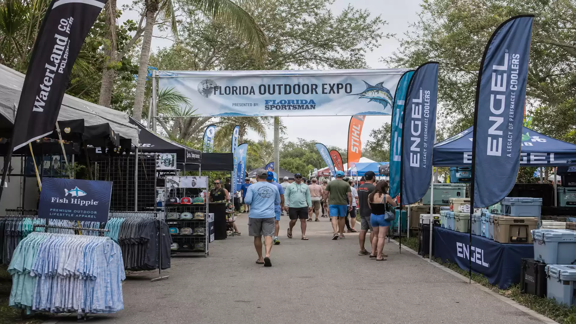 Outdoor expo in Florida with several booths and visitors. Banner with 'Florida Outdoor Expo' and shark graphic. Booths with fishing gear and clothing.