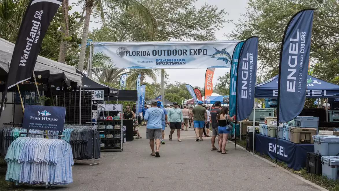 Outdoor expo in Florida with several booths and visitors. Banner with 'Florida Outdoor Expo' and shark graphic. Booths with fishing gear and clothing.
