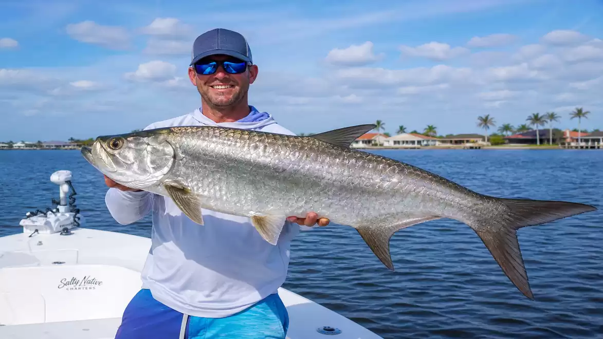Angler holds a large silver fish on a boat with a water landscape, houses, and palm trees in the background.