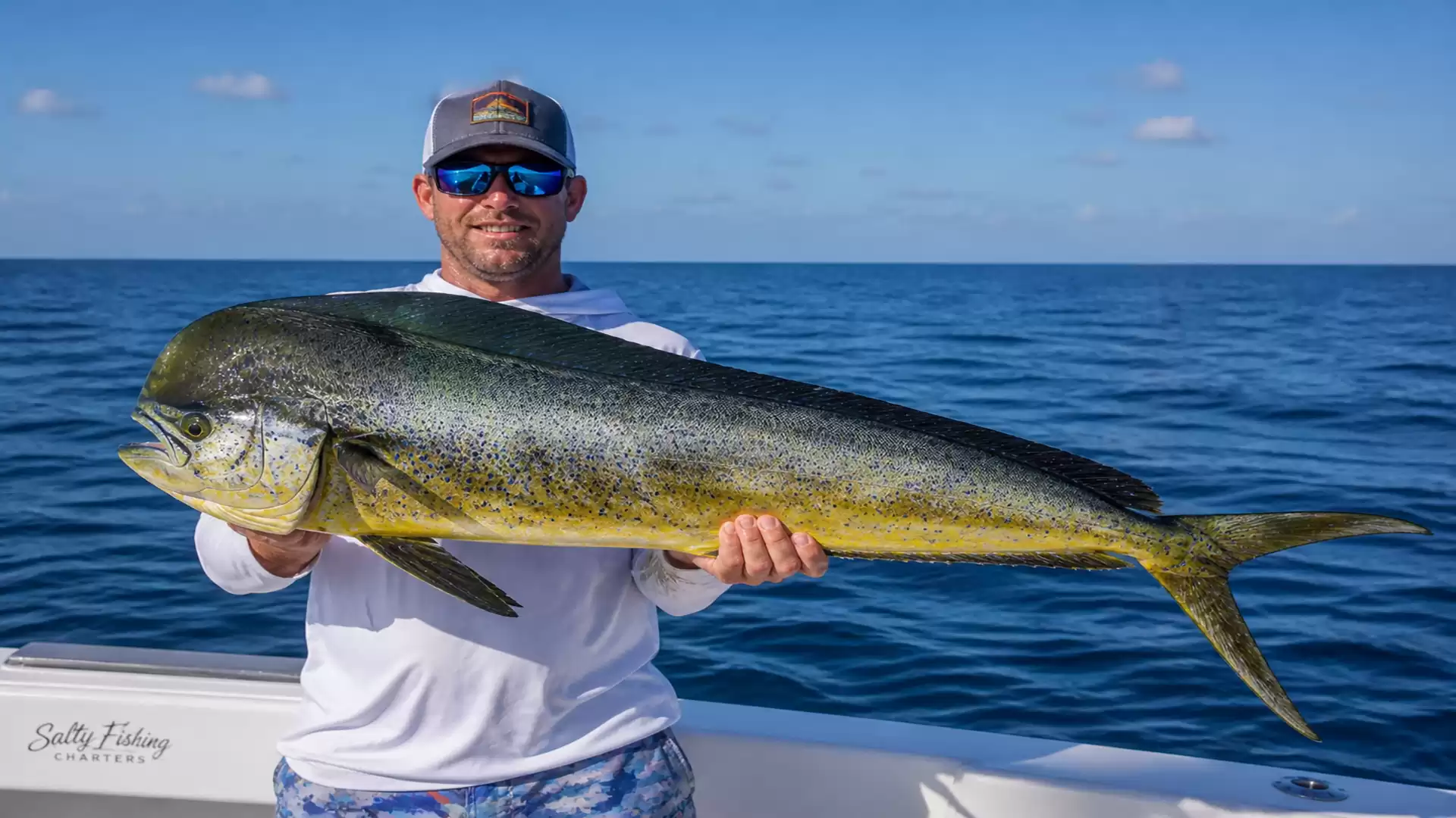 An angler holds a large dolphinfish (mahi-mahi) on a boat, with a blue sky and calm sea in the background.