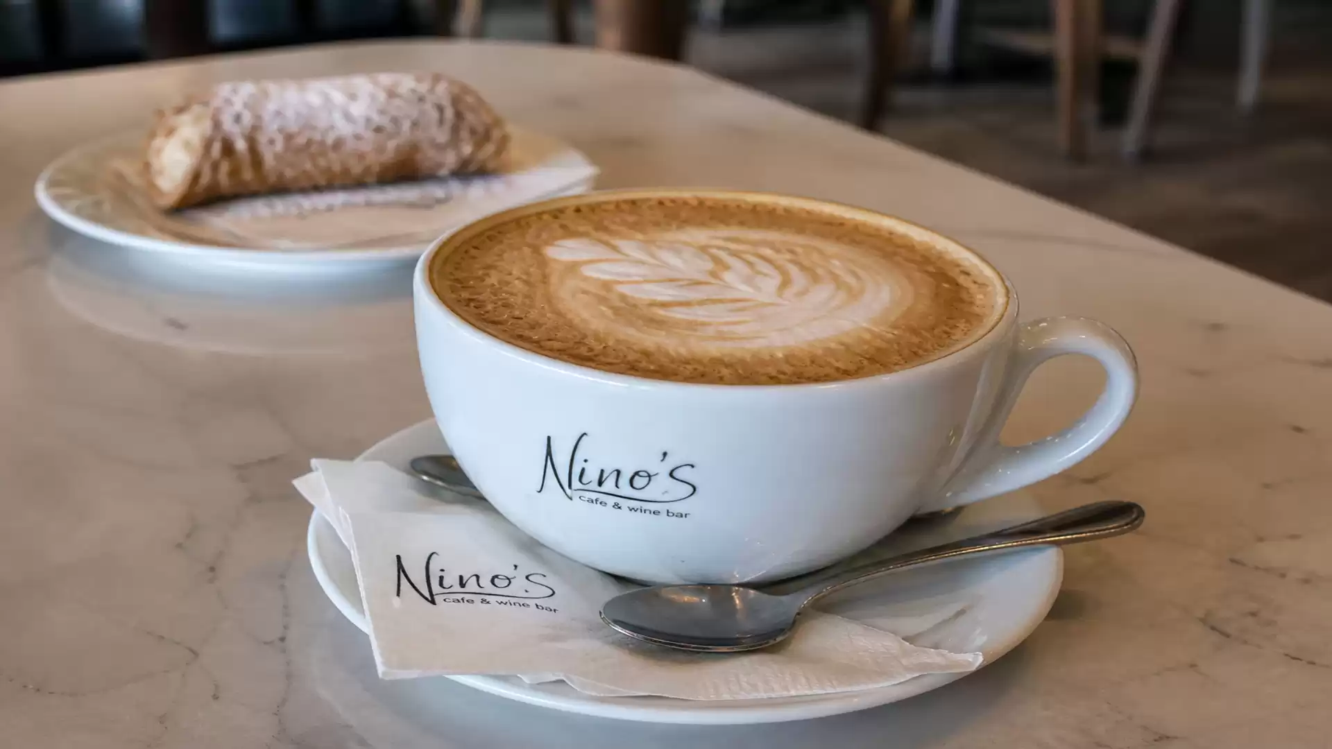 A cappuccino on a marble table in a café, with a sweet pastry on a plate in the background.